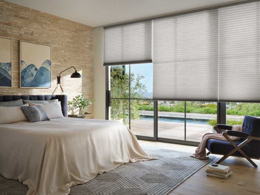 Alustra duette honeycomb automated shades covering floor to ceiling black trim windows in a bedroom. The windows are on the right side of the bed that has two modern paintings above it. The view outside is to a green backyard.